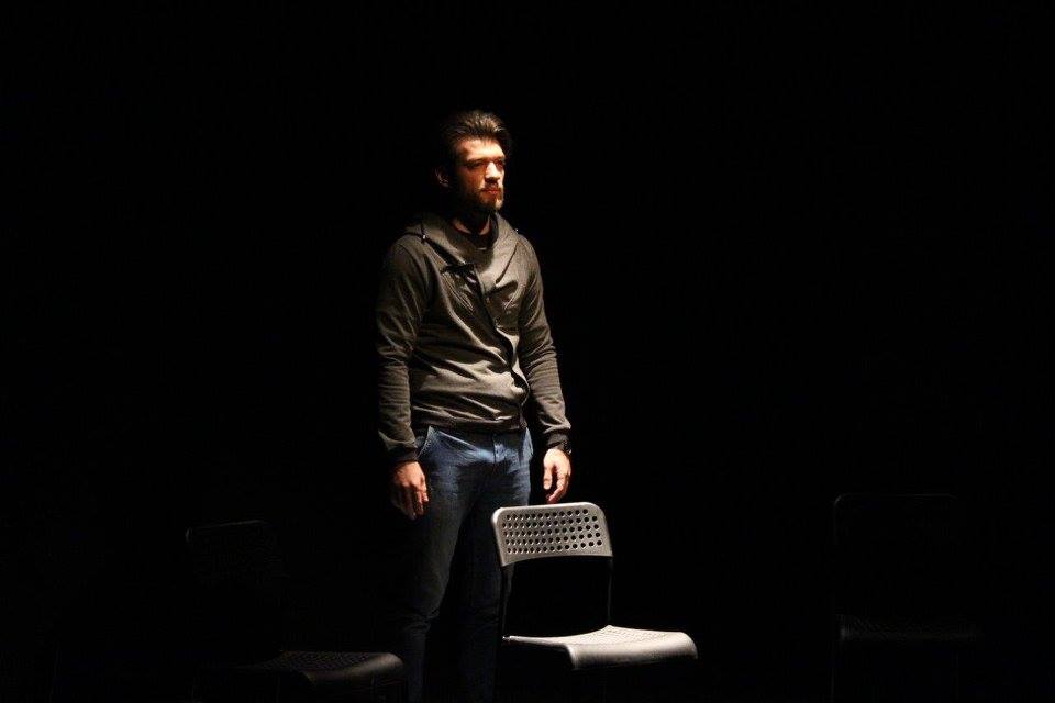 Man standing behind a chair on stage in the dark with a spotlight on him